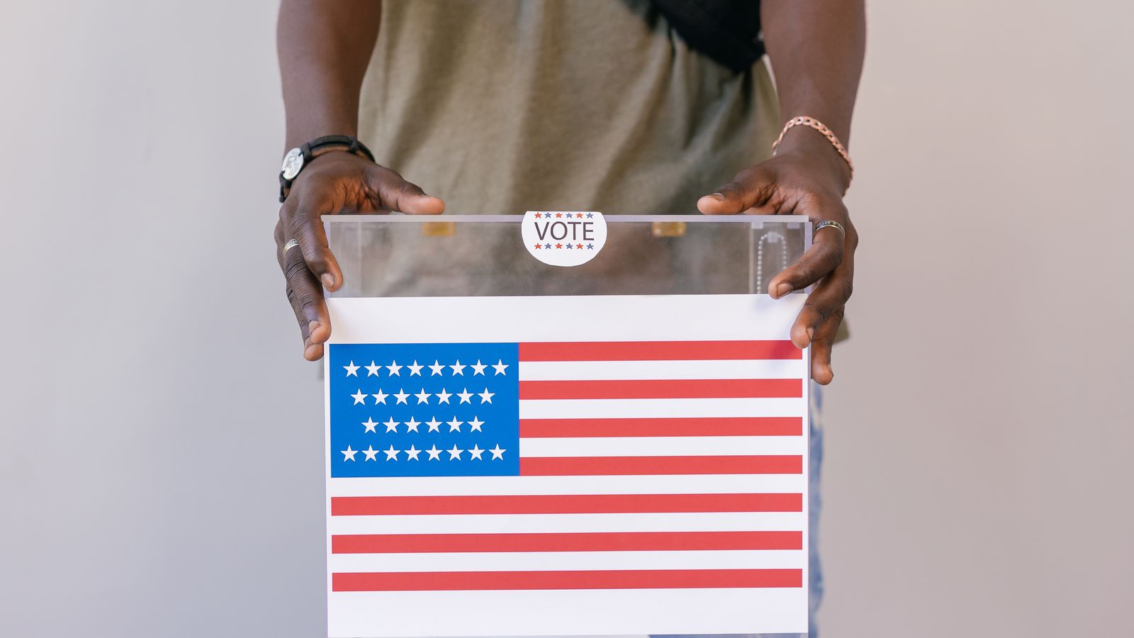 Hands placing a paper ballot into a clear ballot box in front of an American flag